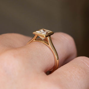A close-up shot of a vintage-inspired princess-cut diamond engagement ring worn on a finger showing the minimalistic rub-over setting.