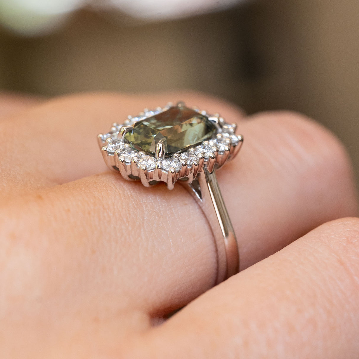 A close-up of the side view of a vintage-inspired, green cushion-cut sapphire and diamond cluster engagement ring in platinum.