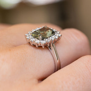 A close-up of the side view of a vintage-inspired, green cushion-cut sapphire and diamond cluster engagement ring in platinum.