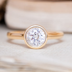 A yellow gold Antique-inspired engagement ring set with a round diamond in a bezel setting, resting on a light textured surface with a softly blurred background.