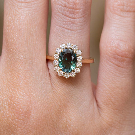 A close-up of a hand wearing a yellow gold engagement ring centred with an alexandrite showing green, blue and purple hues with a halo of round brilliant diamonds. 
