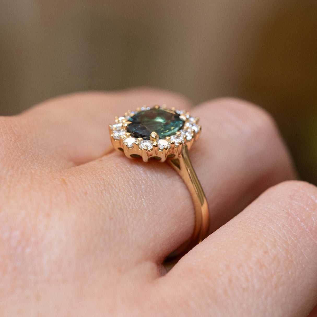 A close-up of a gold engagement ring with an oval green-blue hued gemstone surrounded by round diamonds, showing the setting from the side profile. 