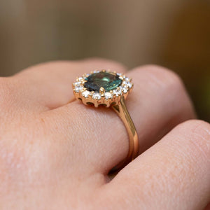 A close-up of a gold engagement ring with an oval green-blue hued gemstone surrounded by round diamonds, showing the setting from the side profile. 