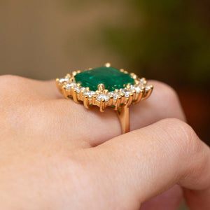 A yellow gold ring with a green emerald  centre stone and diamond halo cluster, shown from the side with a claw-set open back. 