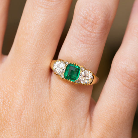 A close-up of an antique, Victorian emerald and diamond three-stone ring set in yellow gold worn on a finger.