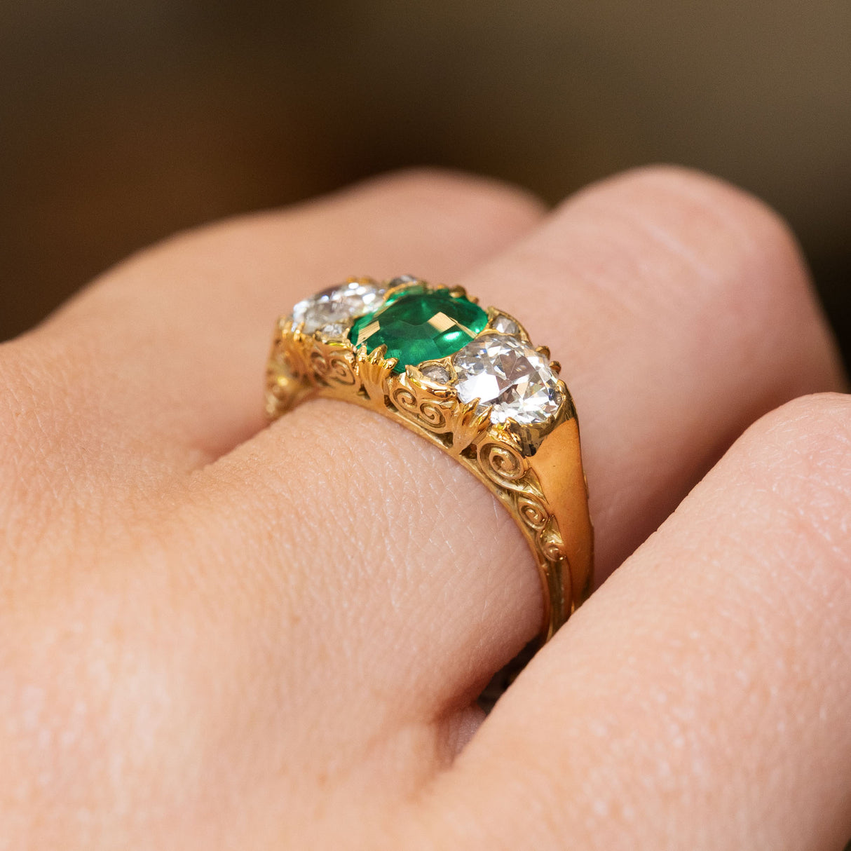 A close-up of an antique green emerald and diamond three-stone ring showing the intricate scroll details engraved into the gallery. 
