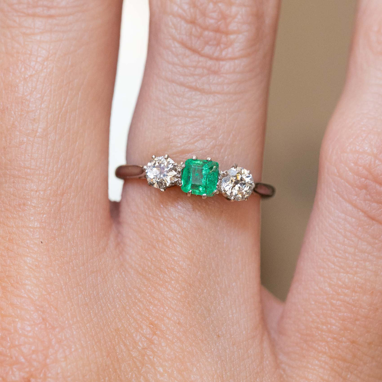 A close-up of a ring worn on a finger, featuring a central green emerald flanked by two old-cut diamonds on a thin white gold band.