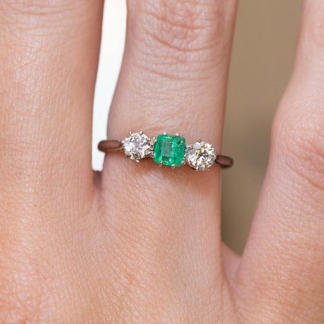 A close-up of a ring worn on a finger, featuring a central green emerald flanked by two old-cut diamonds on a thin white gold band.