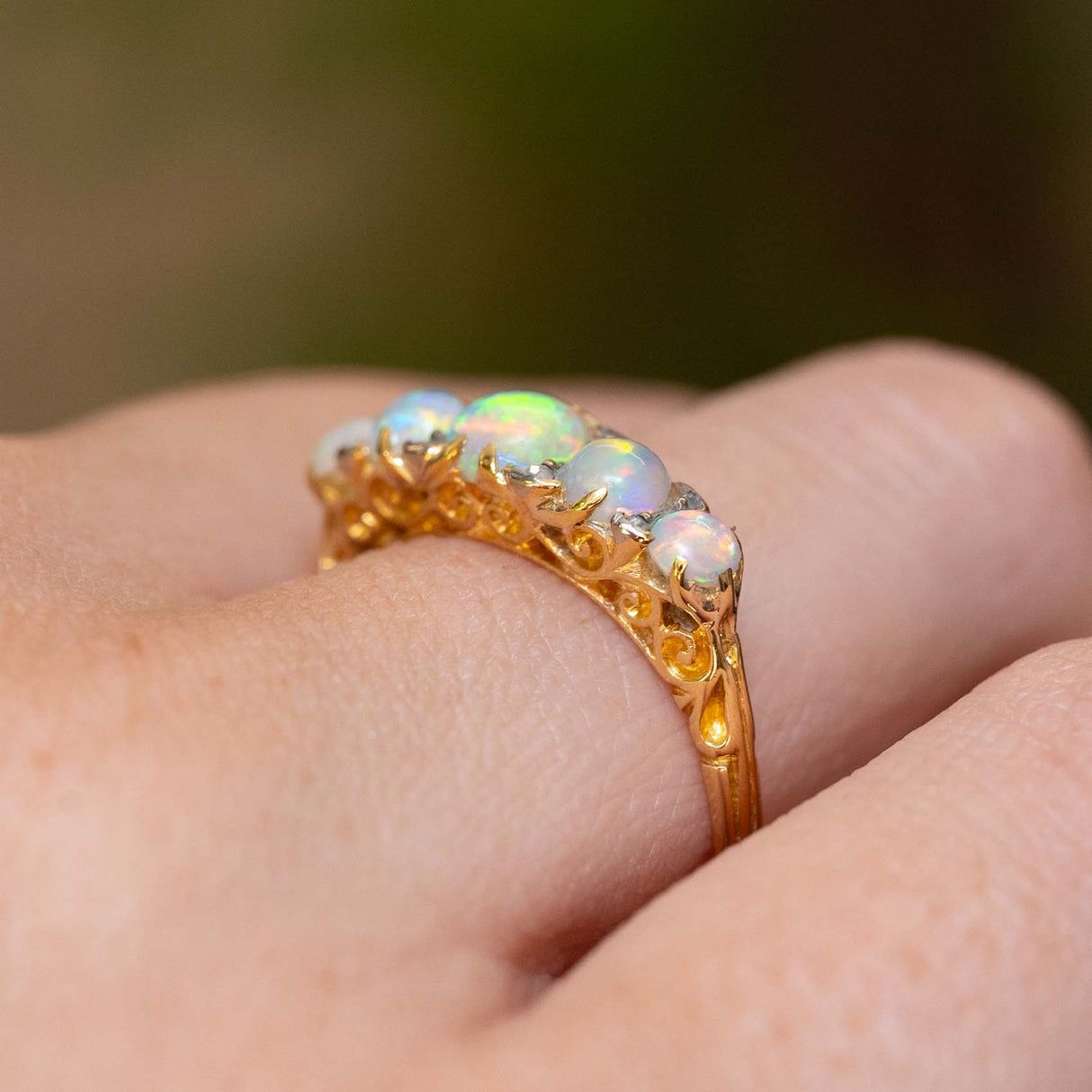 A gold ring with five round iridescent opal gemstones, worn on a person's finger showing the engraved scroll details in the gallery.