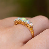 A gold ring with five round iridescent opal gemstones, worn on a person's finger showing the engraved scroll details in the gallery.