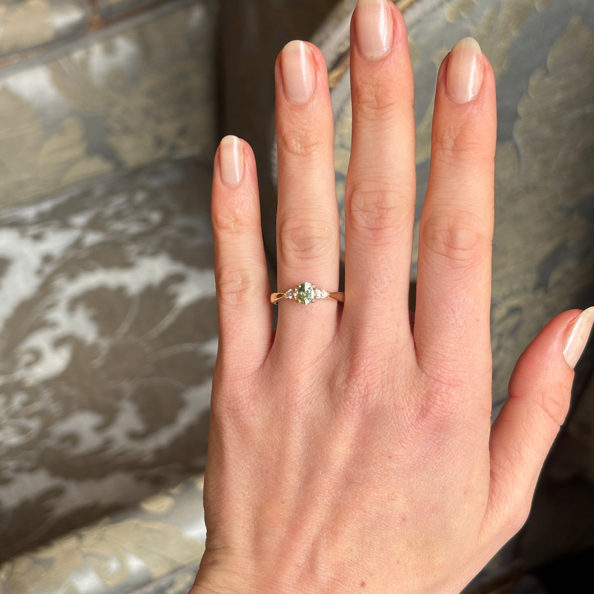 Just-under-1ct green sapphire and diamond three-stone engagement ring worn on the hand, showing the depth of color in a room setting.