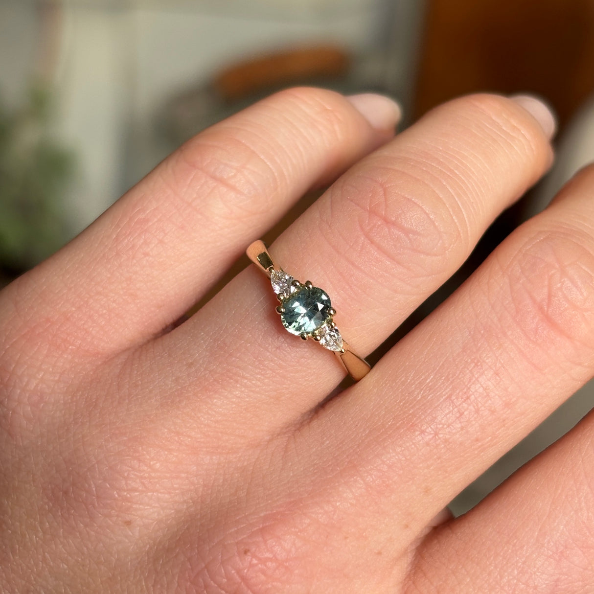 An oval-cut teal sapphire and diamond three-stone engagement ring set in yellow gold, displayed on the engagement finger.