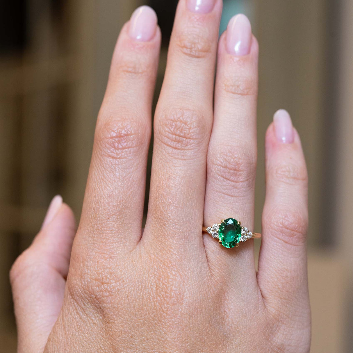 A vintage-inspired oval-cut emerald and diamond trefoil engagement ring worn on the hand.