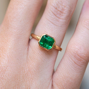 An antique-inspired emerald-cut emerald solitaire engagement ring in 14ct yellow gold worn on the finger.