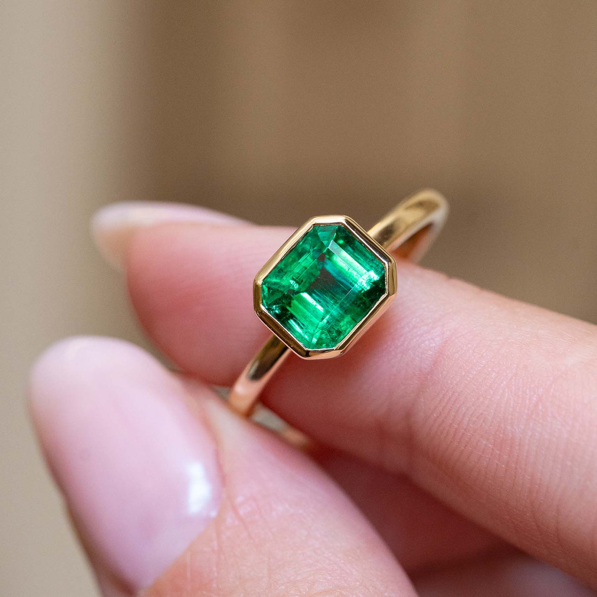 An antique-inspired emerald-cut emerald solitaire engagement ring held between the fingers.