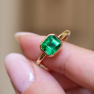 An antique-inspired emerald-cut emerald solitaire engagement ring held between the fingers.