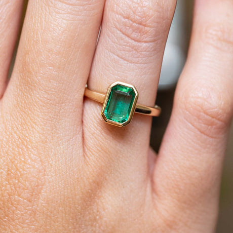 Emerald-cut emerald solitaire engagement ring in yellow gold with an antique-inspired design, worn on the finger.