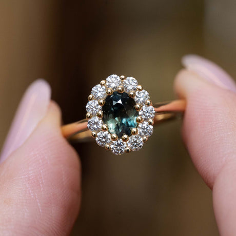 Oval deep green sapphire framed by a sparkling diamond cluster in an antique-inspired yellow gold engagement ring.
