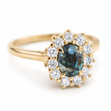 A vintage-inspired, blue-green sapphire and diamond halo engagement ring in 14ct yellow gold.