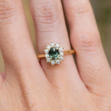 An antique-inspired teal sapphire and diamond cluster engagement ring worn on the finger.