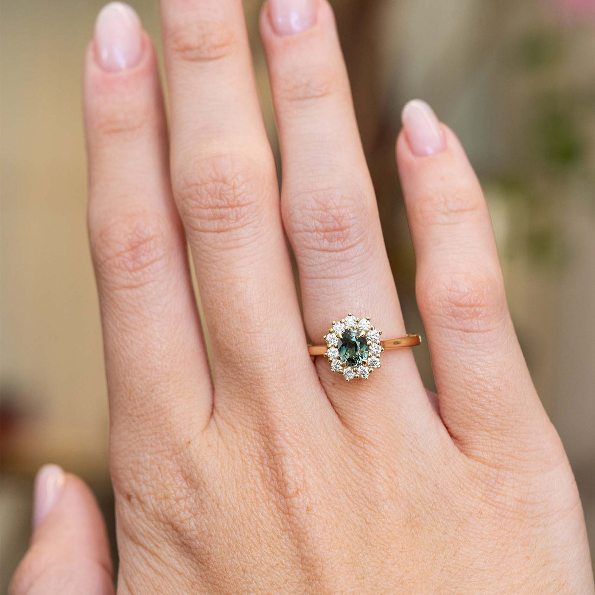 An antique-inspired teal sapphire and diamond halo engagement ring in 14ct yellow gold worn on the hand.