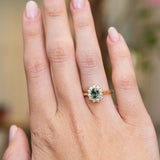 An antique-inspired teal sapphire and diamond halo engagement ring in 14ct yellow gold worn on the hand.
