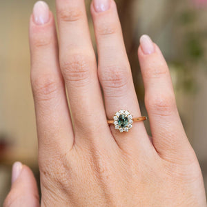 An antique-inspired teal sapphire and diamond halo engagement ring in 14ct yellow gold worn on the hand.
