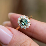 An antique-inspired teal sapphire and diamond halo ring held between the fingers.