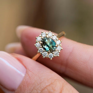 An antique-inspired teal sapphire and diamond halo ring held between the fingers.