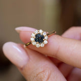An antique-inspired dark teal sapphire and diamond cluster engagement ring held between the fingers.