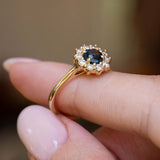 Side detail of an antique-inspired dark teal sapphire and diamond cluster engagement ring held between the fingers.