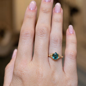 A hand wearing a gold ring with a pear-cut teal tourmaline, set against a softly blurred warm brown background.