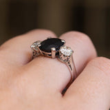 A close-up of a white gold ring with a large dark blue sapphire flanked by a round diamond on either side.