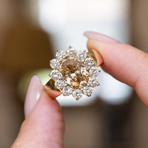 A close-up of a vintage gold ring with a large brown diamond centre stone and a halo diamonds, held between two fingers against a softly blurred warm background.