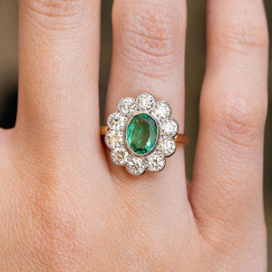 A close-up of a hand wearing a vintage  yellow and white gold ring with a green emerald gemstone and diamond cluster ring.