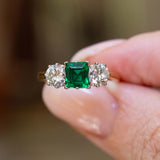 A vintage 1960s emerald and diamond three-stone ring held between the fingers.