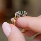 Gallery detail of a vintage 1960s emerald and diamond three-stone ring held between the fingers.