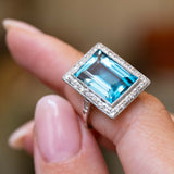 An antique Art Deco aquamarine and diamond cocktail ring in platinum held between the fingers.