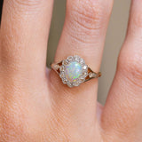 A close-up of a ring worn on a finger, featuring an oval iridescent opal center stone surrounded by a halo of old-cut diamonds.