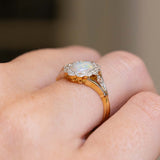 A close-up of a gold and platinum ring with a central iridescent opal surrounded by old-cut diamonds, worn on a person's finger.