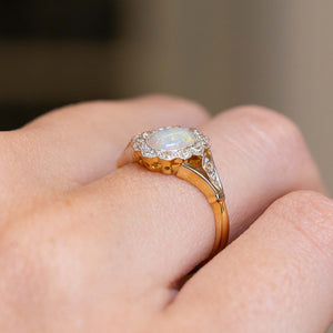 A close-up of a gold and platinum ring with a central iridescent opal surrounded by old-cut diamonds, worn on a person's finger.