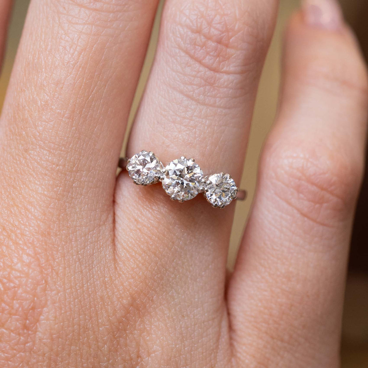 A close-up of a hand wearing a three-stone diamond ring. The ring features three round old-cut diamonds set in a platinum band.