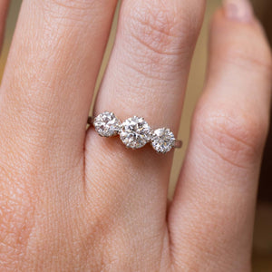 A close-up of a hand wearing a three-stone diamond ring. The ring features three round old-cut diamonds set in a platinum band.