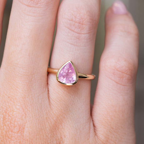 A close-up photo of an Antique-inspired, yellow gold engagement ring featuring a baby Pink sapphire, pear-cut and bezel set.