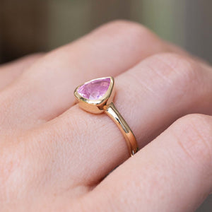 A detailed photo of a yellow gold ring with a drop shaped light pink sapphire drop-cut and rub-over set on an antique inspired solitaire engagement ring, shown from an angle to show how it fits on the finger. 