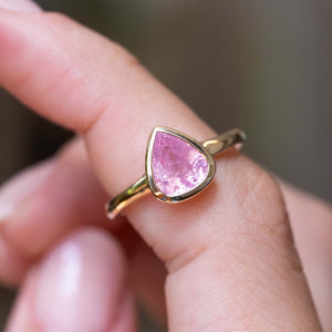 A close-up picture of a yellow gold solitaire engagement ring with a teardrop-shaped pink sapphire that is antique-inspired, placed on a finger.
