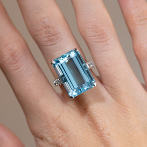 Close-up of a vintage 1970s aquamarine cocktail ring worn on a finger, featuring a large emerald-cut blue aquamarine flanked by baguette-cut diamonds in white gold.