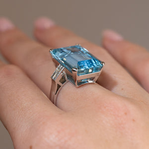 Side profile of a vintage 1970s aquamarine cocktail ring worn on a finger, revealing its raised open setting, diamond-set shoulders and white gold band.