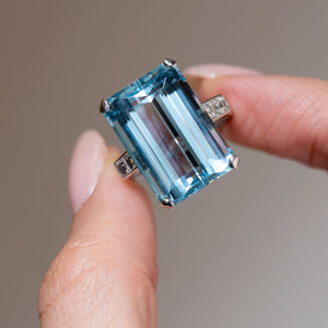 Detailed close-up of a vintage 1970s aquamarine and diamond cocktail ring held between fingers, highlighting the aquamarine's crisp step-cut facets and four-claw setting.