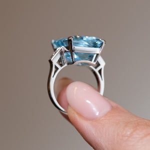 Side profile of a vintage 1970s aquamarine and diamond cocktail ring held between fingers, showing the elevated gemstone, architectural open gallery and geometric white gold shoulders.